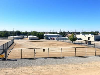 Mixed Use Land for Sale in Ajman Industrial, Ajman - A_vacant_industrial_plot_in_an_urban_area_featuring_open_land_ready_for_development. _The_scene_includes_a_flat_expanse_of_dirt_and_gravel_surrounded_by_chain_link_fencing. _Nearby_there_are_scattered_industrial_buildi. jpeg
