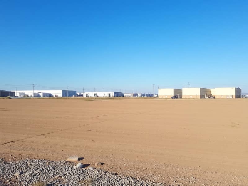 2 A_vast_empty_industrial_plot_of_land_covered_with_dirt_and_gravel_ready_for_future_construction. _In_the_background_there_are_several_nearby_industrial_buildings_and_warehouses_under_a_clear_blue_sky. _Utility_poles_ca. jpeg