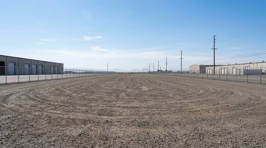 Plot zum zum Verkauf in Emirates Industrial City, Sharjah - A_very_large_empty_industrial_photo_landscape_in_wide_angle_perspective_a_clean_vacant_industrial_yard_with_a_wide_dirt_gravel_ground_distant_warehouse_buildings_on_the_left_and_right_a_tall_chain_link_fence_enclosing. png
