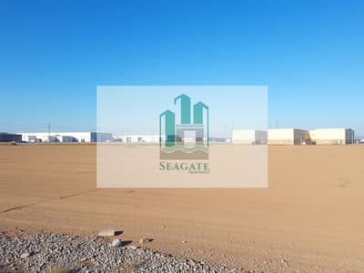 Mixed Use Land te Koop in Al Alia, Ajman - A_vast_empty_industrial_plot_of_land_covered_with_dirt_and_gravel_ready_for_future_construction. _In_the_background_there_are_several_nearby_industrial_buildings_and_warehouses_under_a_clear_blue_sky. _Utility_poles_ca. jpeg