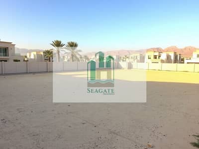 Plot zum Verkauf in Al Jurf, Ajman - A_vacant_residential_plot_in_Al_Jurf_UAE_characterized_by_a_flat_sandy_terrain_bordered_by_modern_villas. _The_plot_is_enclosed_with_a_fence_and_has_a_backdrop_of_palm_trees_and_distant_mountains. _The_sun_casts_a_warm. jpeg