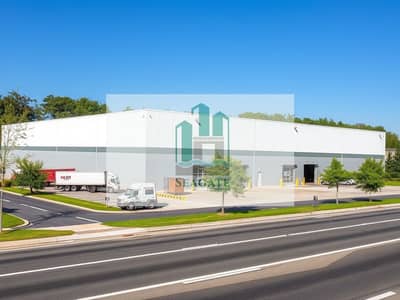 阿联酋工业城， 夏尔迦 仓库待租 - A_warehouse_exterior_located_near_a_roadside. _The_scene_includes_a_large_modern_warehouse_building_with_a_flat_roof_surrounded_by_a_parking_area_and_a_loading_dock. _There_are_delivery_trucks_parked_nearby_and_trees. jpeg