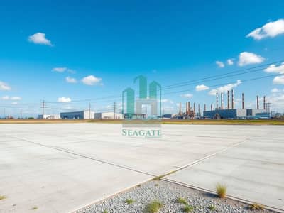 ارض صناعية  للايجار في صناعية الصجعة، الشارقة - A_vast_industrial_land_plot_showcasing_an_expansive_area_of_flat_concrete_and_gravel. _The_surroundings_consist_of_several_large_warehouses_and_factories_with_smokestacks_in_the_background. _The_sky_is_bright_blue_with. jpeg