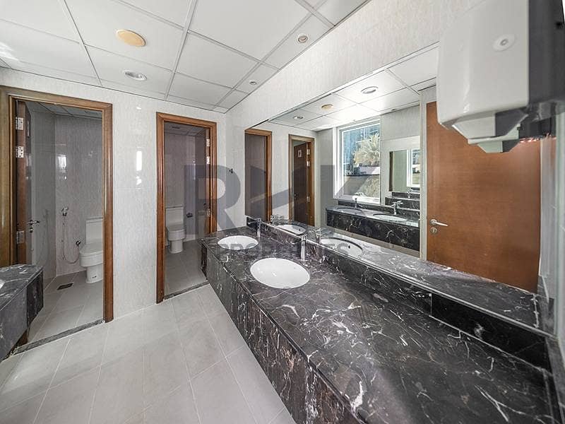 26 Fitted office restroom in Ontario Tower Business Bay with dark marble countertop, twin basins, WC cubicles and a large mirror
